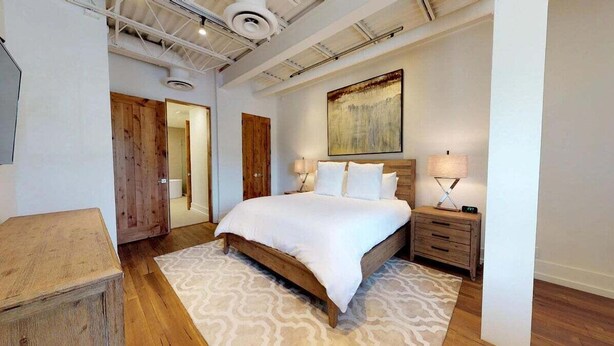 A bedroom with a wooden bed, bedside lamps, a wooden nightstand, a patterned rug, and a large abstract painting on the wall.