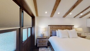 Een slaapkamer met een houten hoofdbord, twee nachtkastjes met lampen, een bed met wit beddengoed en een raam met zonwering.