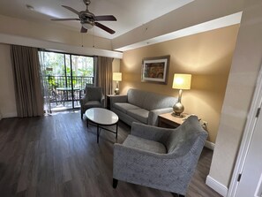 A hotel room with a ceiling fan, a sofa, a chair, a small table, and a lamp. There is a window with curtains and a view of greenery outside.