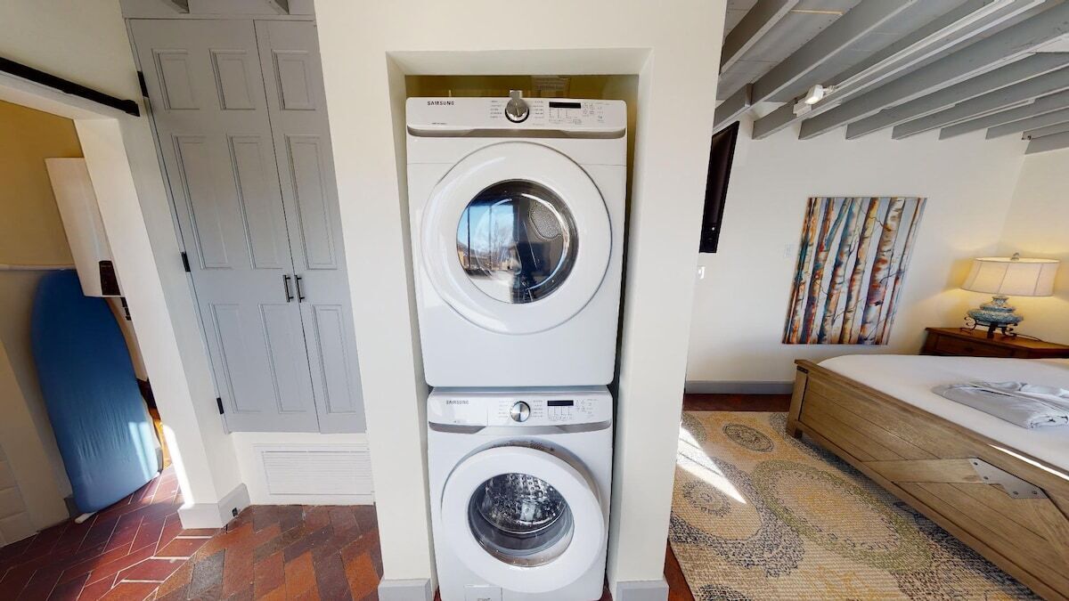 Une buanderie avec des machines à laver et des sèche-linge superposés, un lit avec une table de chevet et un tableau accroché au mur.