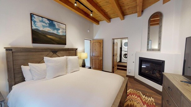 A bedroom with a bed, a fireplace, a mirror, a painting, and a door leading to a laundry area.