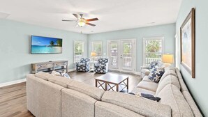 A living room with a beige sectional sofa, a coffee table, and patterned armchairs. There is a ceiling fan, a painting of a beach scene, and a framed picture on the wall.