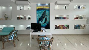 Una sala de estar moderna con un cuadro central, un escritorio blanco con una computadora y sillas de mimbre. Estanterías con libros y objetos decorativos bordean las paredes.