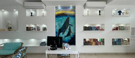 A modern living room with a central painting, a white desk with a computer, and wicker chairs. Shelves with books and decorative items line the walls.
