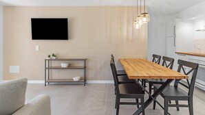 Un comedor moderno con una mesa de madera, sillas negras, un televisor en la pared y un estante pequeño con plantas.