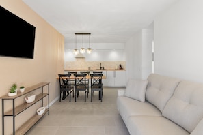 A modern living room with a sofa, a dining area with chairs, a TV mounted on the wall, and a kitchen in the background.
