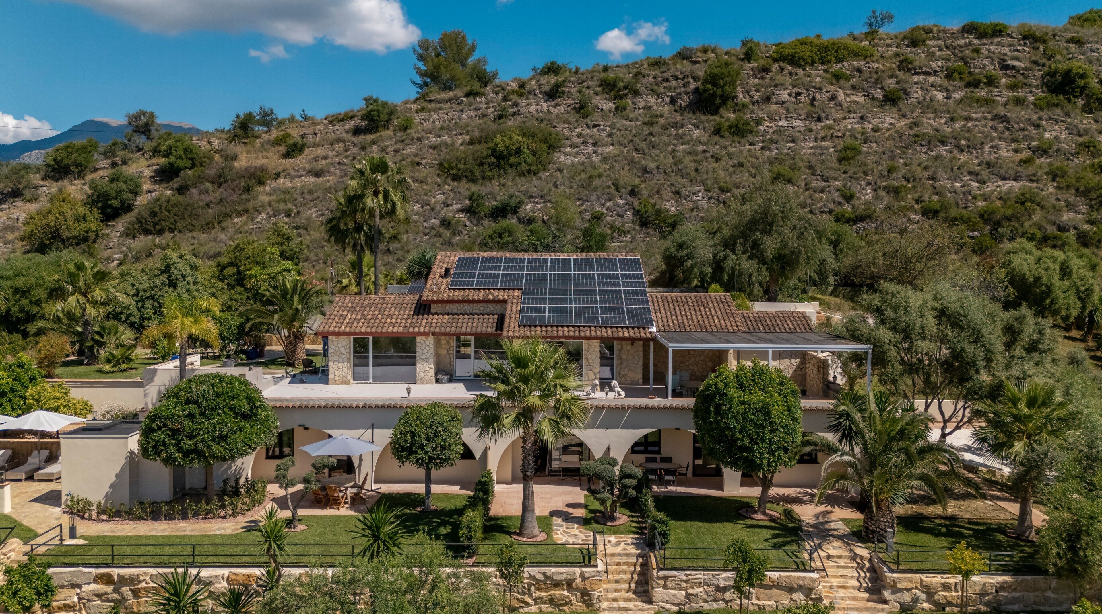 A house with a solar panel roof, stone walls, and a landscaped garden with palm trees.