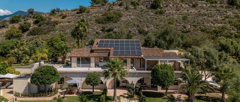 A house with a solar panel roof, stone walls, and a landscaped garden with palm trees.