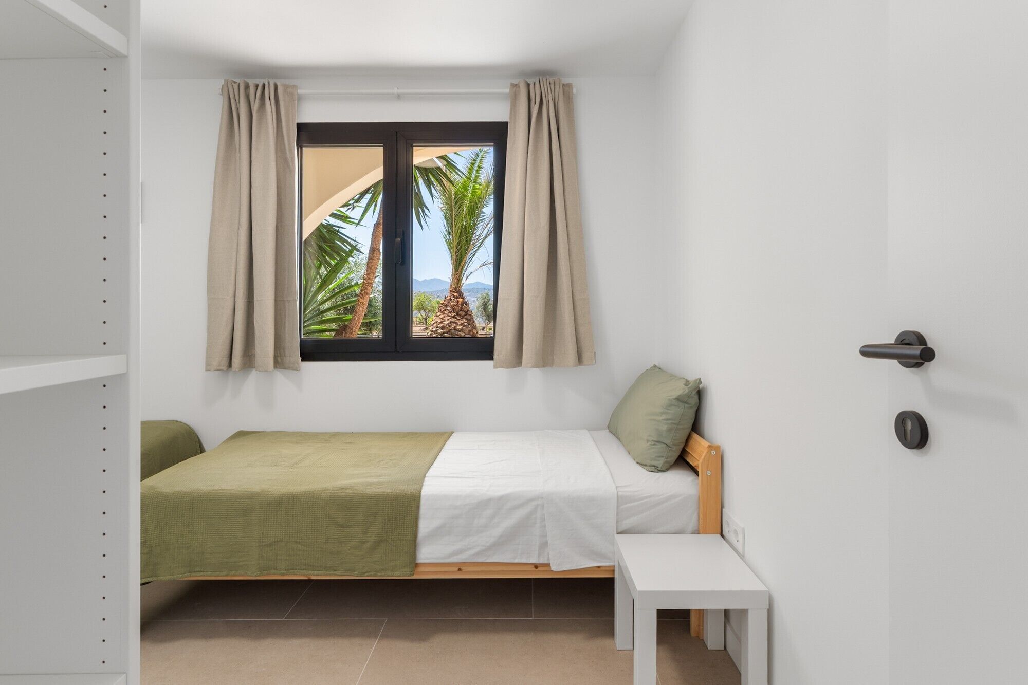 A small, single-bed room with a window, a bedside table, and a door.