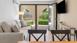 Una sala de estar moderna con un sofá, una mesa, sillas, un televisor montado en la pared y un ventanal que da a un espacio exterior con vegetación.