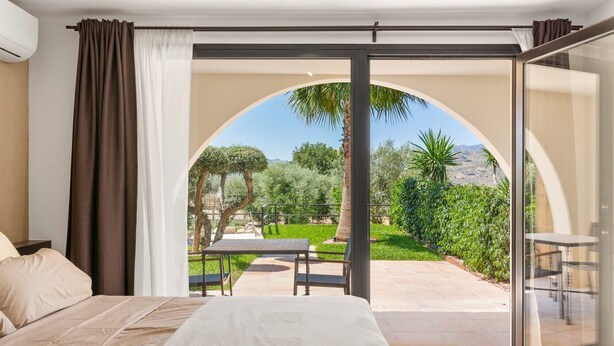 Un dormitorio con una cama, vista a un patio a través de un arco, con una mesa y sillas, y una palmera.