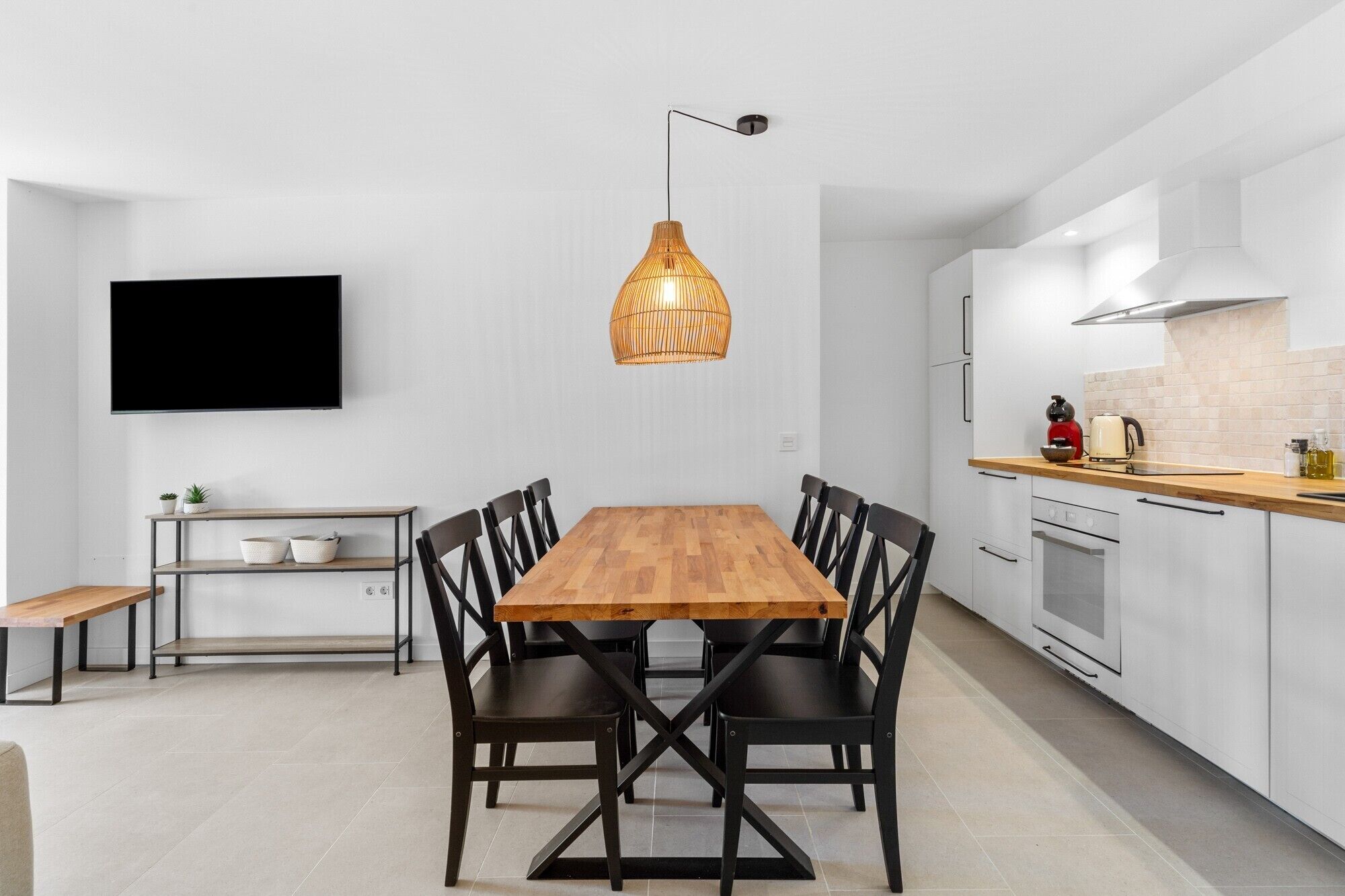 Une cuisine moderne avec des meubles blancs, une table à manger en bois, des chaises noires, un luminaire suspendu et un téléviseur à écran plat fixé au mur.