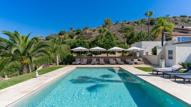 Piscina con sillones de descanso y sombrillas, rodeada de vegetación y una ladera.
