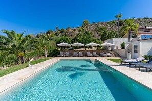 Piscina con sillones de descanso y sombrillas, rodeada de vegetación y una ladera.