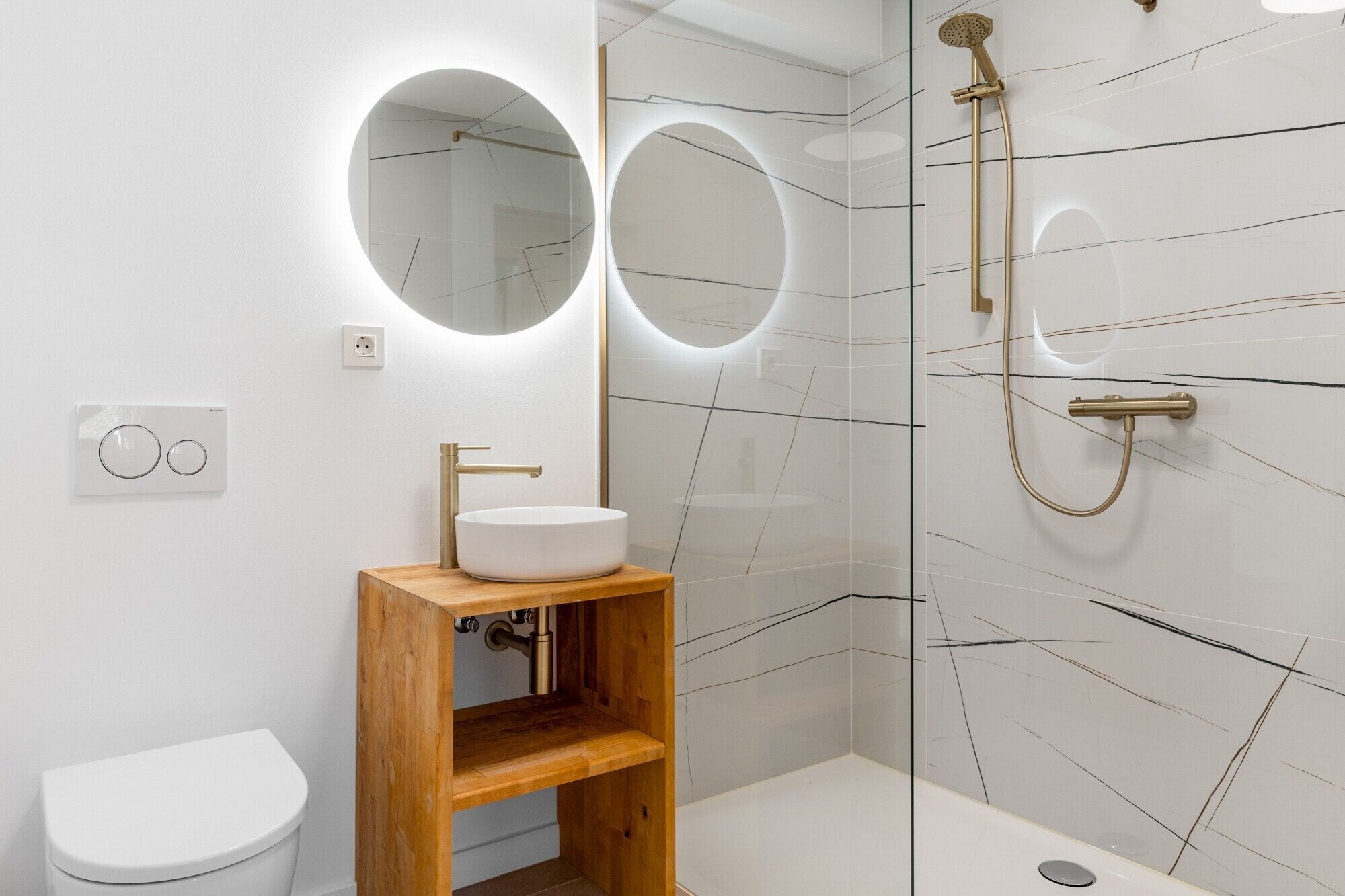Un baño moderno con lavamanos blanco, tocador de madera y zona de ducha con mampara de vidrio.