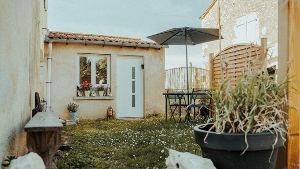 Eine rustikale Terrasse im Freien mit einem Holztisch, Stühlen, einem Topfpflanzen und einer weißen Tür.