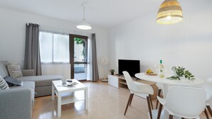 A modern living room with a sofa, dining table, chairs, a flat-screen TV, and a ceiling light fixture. - Oasis Duna Surfhouse Corralejo Vacation Rental with Shared Pool, Private Terrace & Wi-Fi (Corralejo)