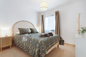 Un lit recouvert d’une couvre-lit à motifs floraux, une lampe de chevet, une table de nuit en bois et une fenêtre avec des rideaux.