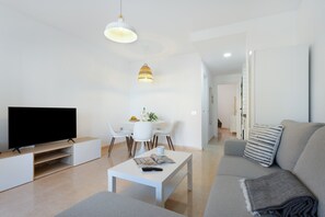Una sala de estar moderna con un televisor de pantalla plana, una mesa de centro blanca, un sofá gris y un comedor con una mesa y sillas blancas.