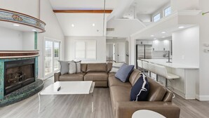A spacious living room with a fireplace, a large sofa, and a kitchen area with white cabinets. - Charming newly updated home in Avalon with endless views of bay and sunset (Avalon)