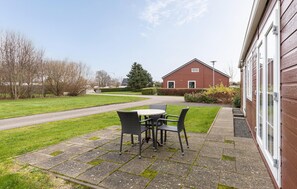 Une terrasse avec une table et des chaises, un bâtiment rouge en arrière-plan et une zone herbeuse avec des arbres.