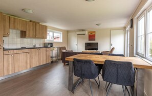Une cuisine moderne avec des meubles en bois, un coin repas avec une table en bois et des chaises noires, un téléviseur et une grande fenêtre qui laisse entrer la lumière naturelle.