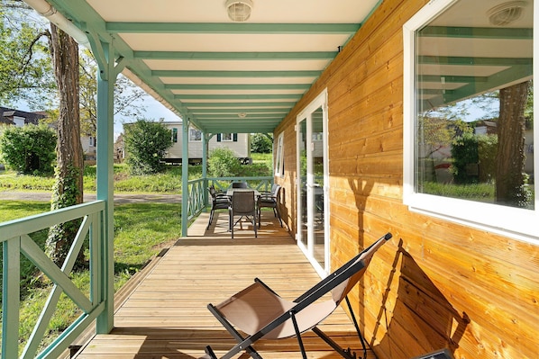 A wooden deck with a table and chairs, a lounge chair, and a covered porch with a railing.
