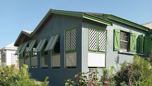 A single-story house with a green roof and shutters, surrounded by a grassy yard and various plants.