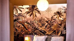 Una cama con un tapiz de pared con motivos tropicales, una lámpara colgante y una mesita de noche con una lámpara.