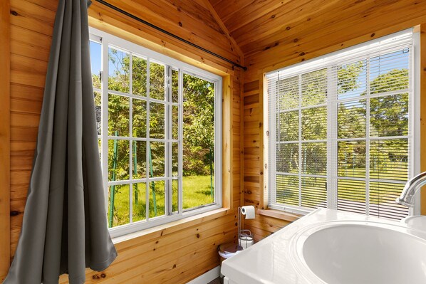 Une salle de bain rustique, aménagée dans une cabane en bois, avec une baignoire, des toilettes et une fenêtre équipée de stores.