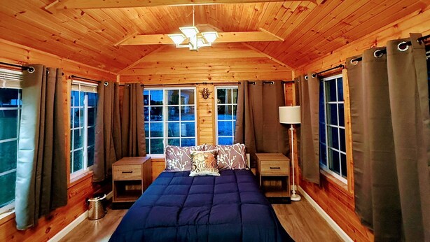 Dormitorio de una cabaña de madera con una cama, mesitas de noche y un ventanal con cortinas.