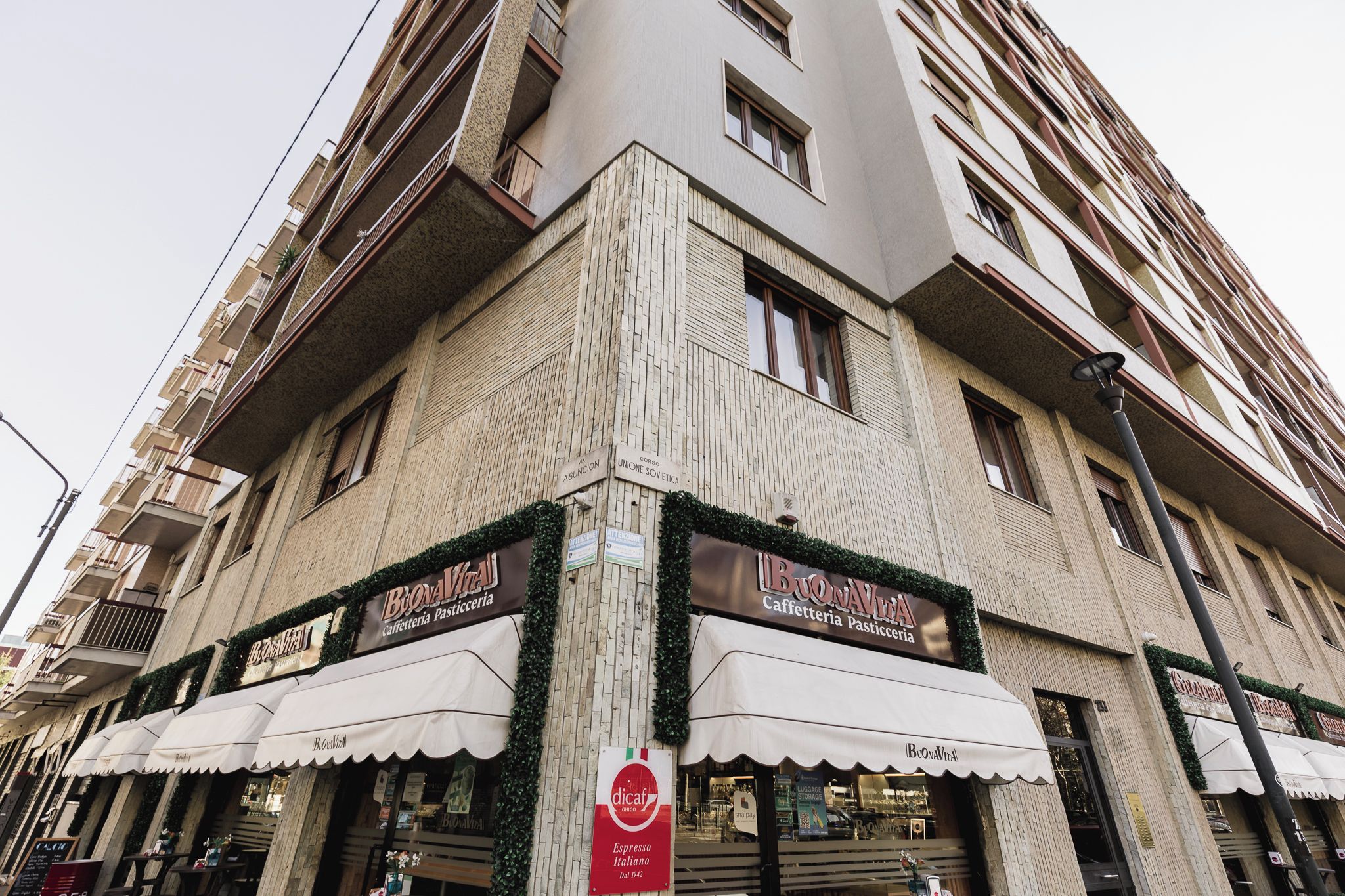 Un edificio ad angolo con vetrine, tra cui un caffè chiamato 'BuonaVita' e un'insegna per 'dicapio' che indica una caffetteria.