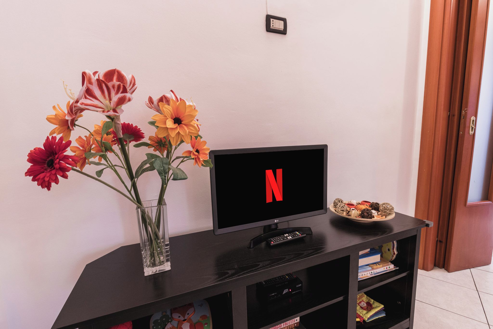 A flat-screen TV with the Netflix logo on a black stand, a vase with flowers, and a plate with snacks.