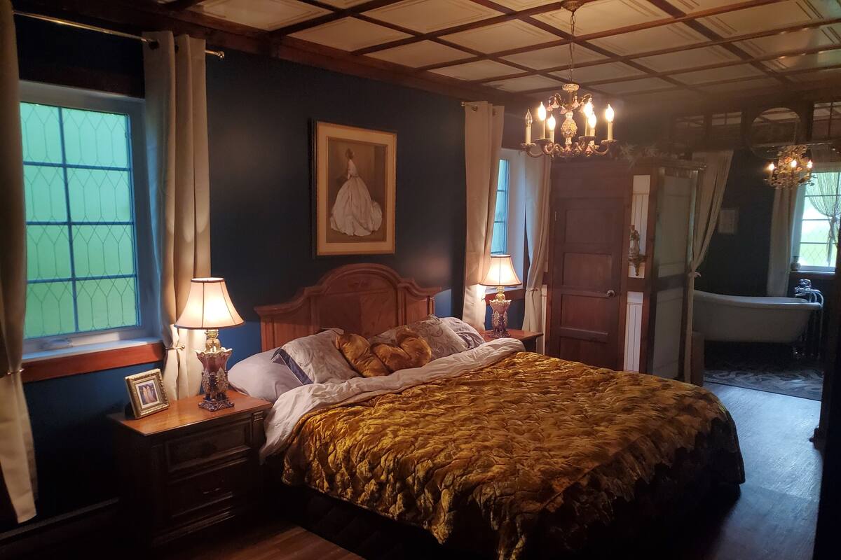 A bedroom with a large bed, two bedside lamps, a wooden headboard, a framed picture on the nightstand, and a chandelier.