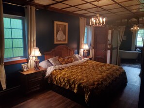 A bedroom with a large bed, two bedside lamps, a wooden headboard, a framed picture on the nightstand, and a chandelier. - Le Manoir aux Secrets (Rawdon)