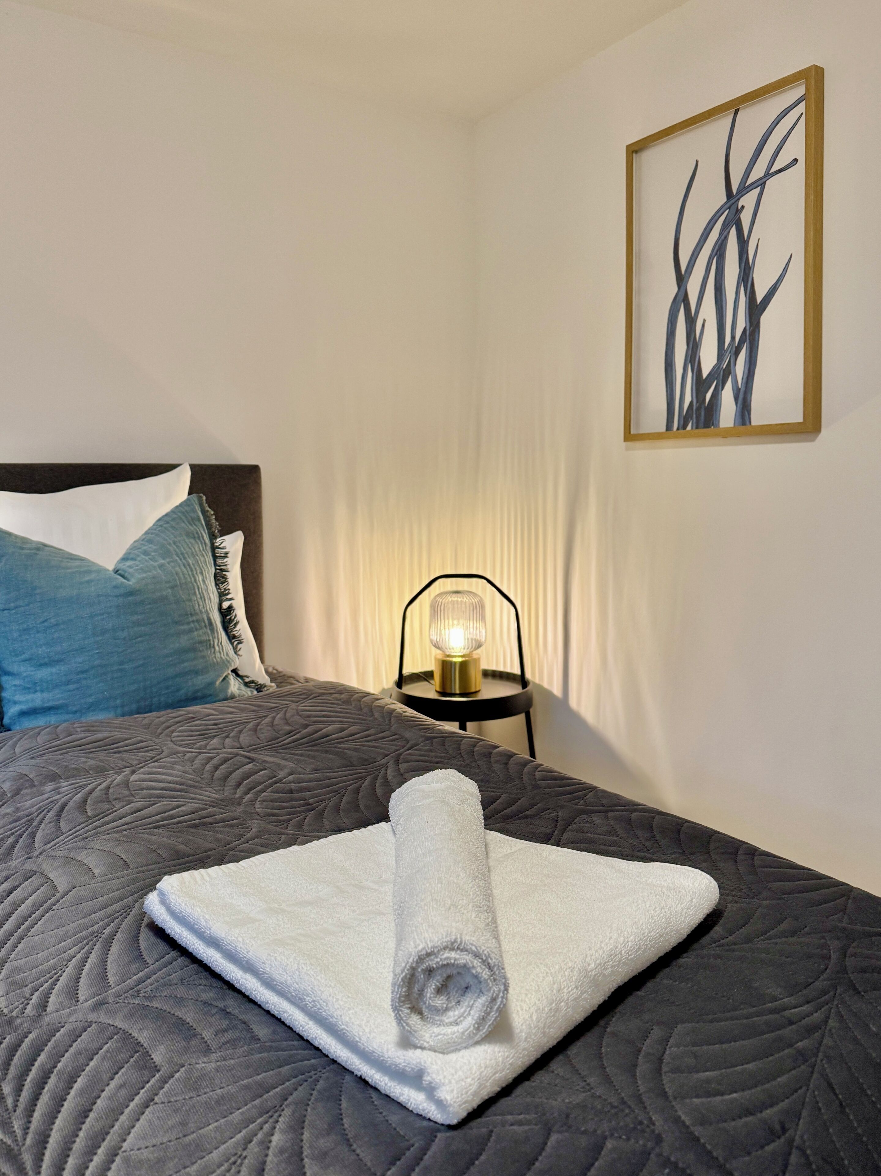 Una cama bien hecha con una toalla doblada, una lámpara de noche y un cuadro abstracto enmarcado en la pared.