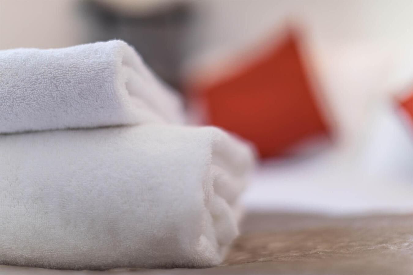 Une serviette blanche, soigneusement pliée, est posée sur une surface en bois, avec un arrière-plan flou où l’on aperçoit un coussin rouge.