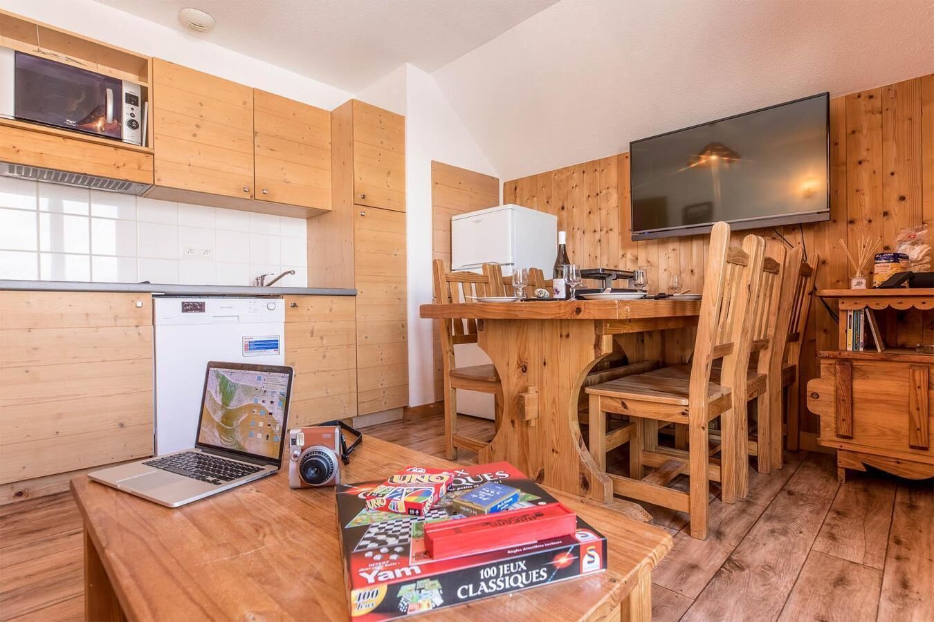 Een compacte keuken met houten kastjes, een magnetron, een koelkast en een eethoek met houten stoelen.
