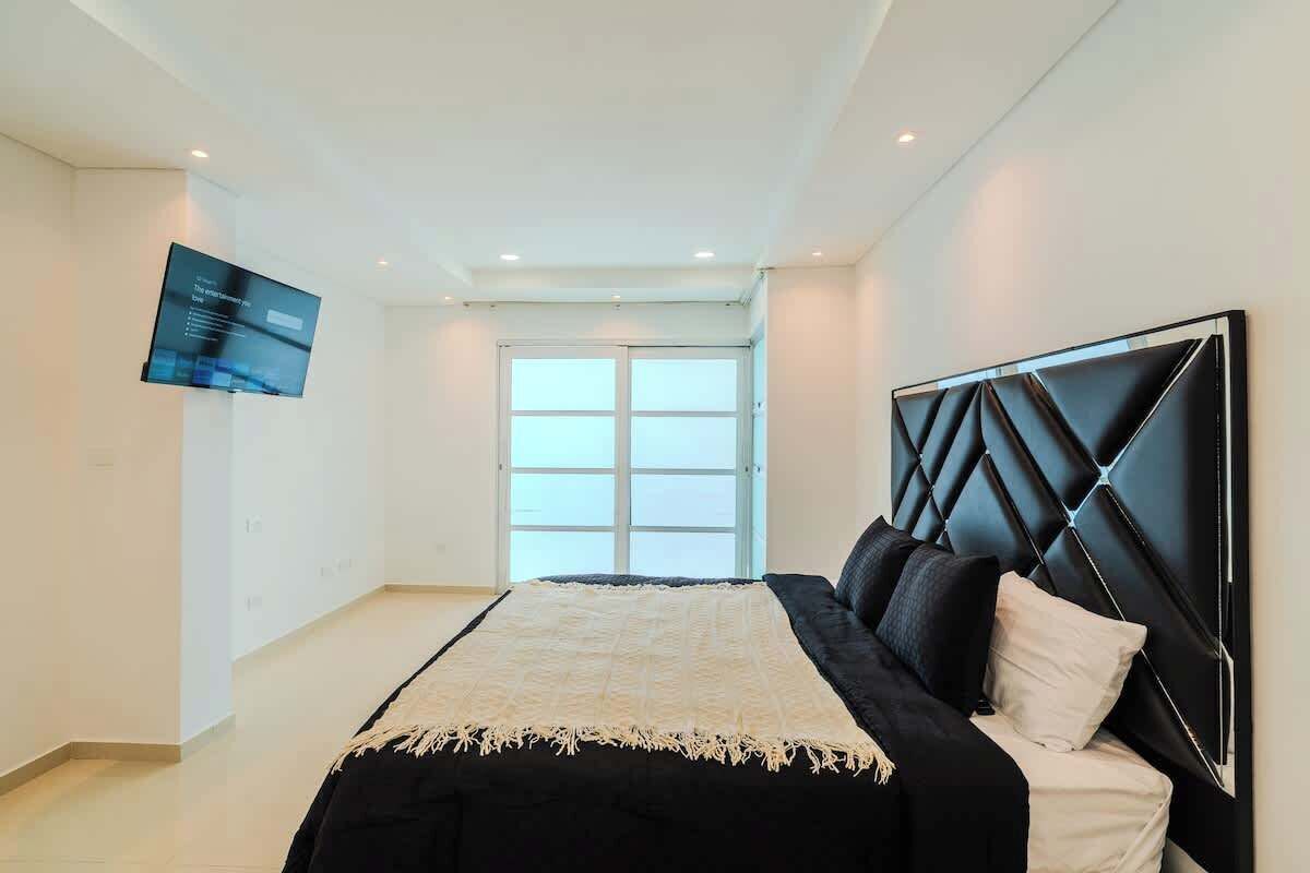 A modern bedroom with a large bed, a flat-screen TV mounted on the wall, and a large window providing natural light.