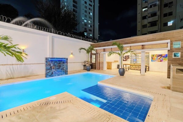 Una piscina con un borde de baldosas, una zona de deck de madera y un edificio con un mural.