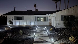 Una casa moderna con un camino de entrada bien iluminado, el número '8308' en la pared y un pequeño jardín con rocas y plantas.