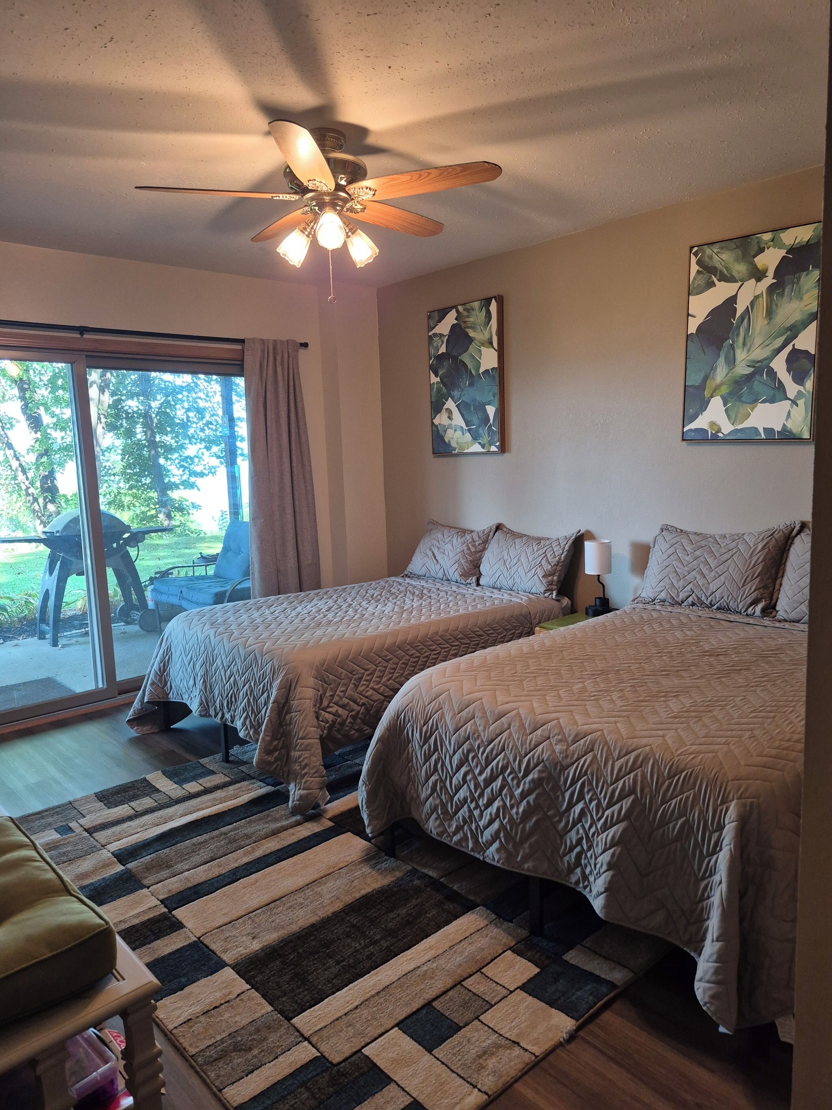 A bedroom with two beds, a ceiling fan, a window with a view of the outdoors, and a door leading to a patio.