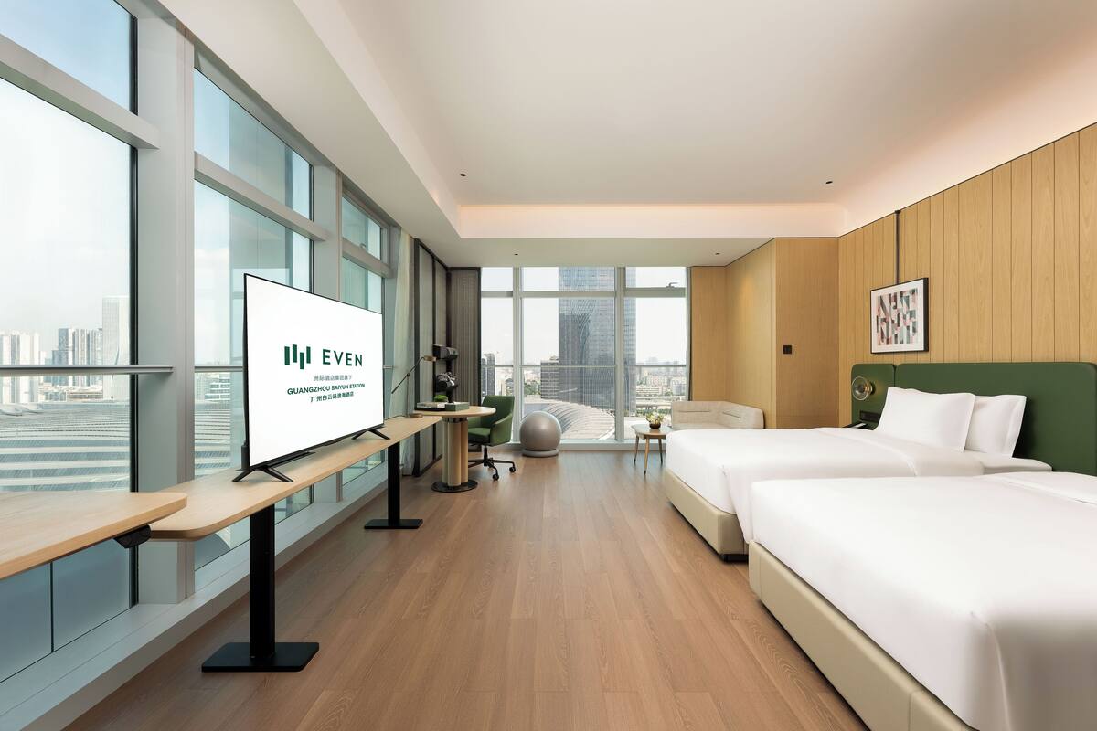 Una habitación de hotel moderna con un ventanal grande, un escritorio, dos camas y una pared verde.
