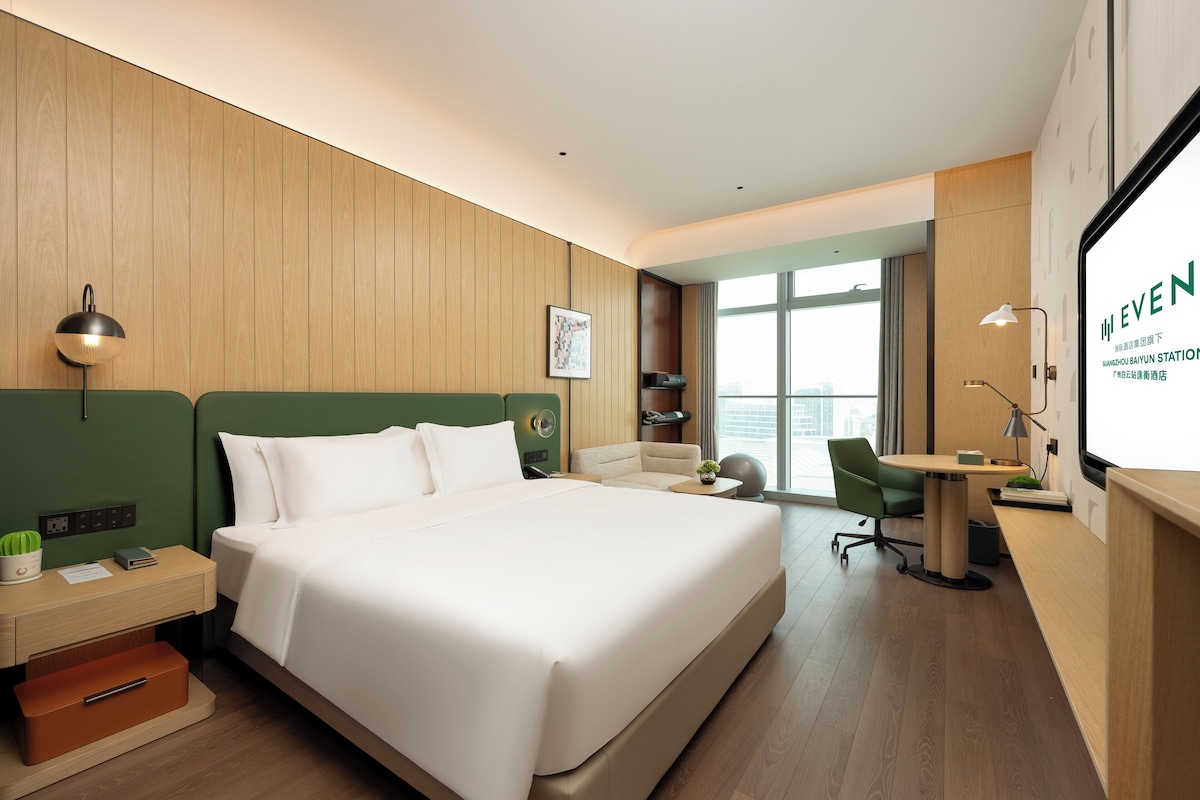 A hotel room with a large bed, a desk, a chair, a nightstand, and a window with a view.