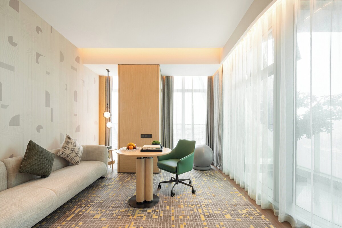 A modern hotel room with a sofa, a green armchair, a small round table with a lamp, and a large window with sheer curtains.