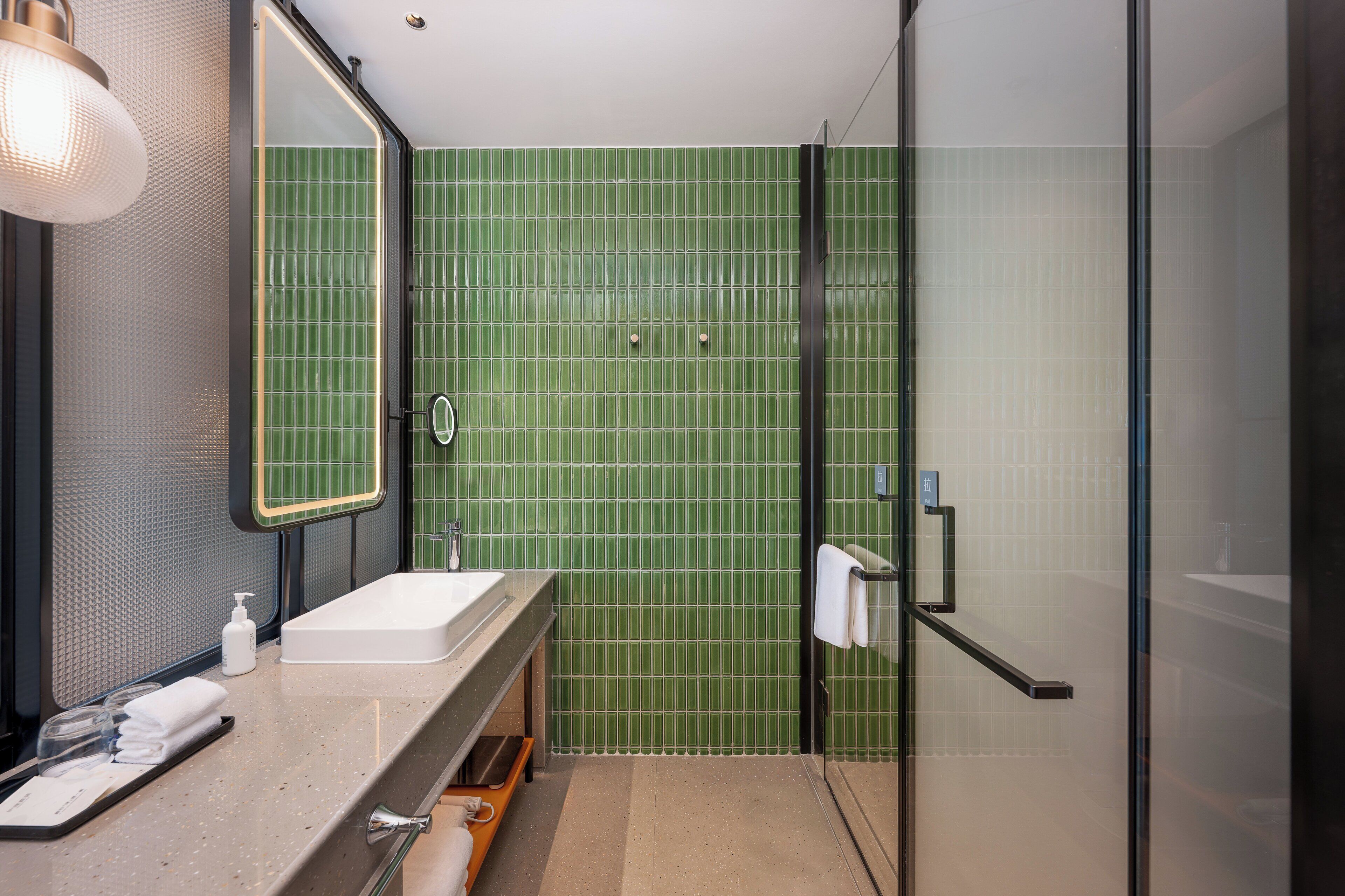 Une salle de bain moderne avec un mur de douche recouvert de carreaux verts, un lavabo blanc et une douche à parois de verre.