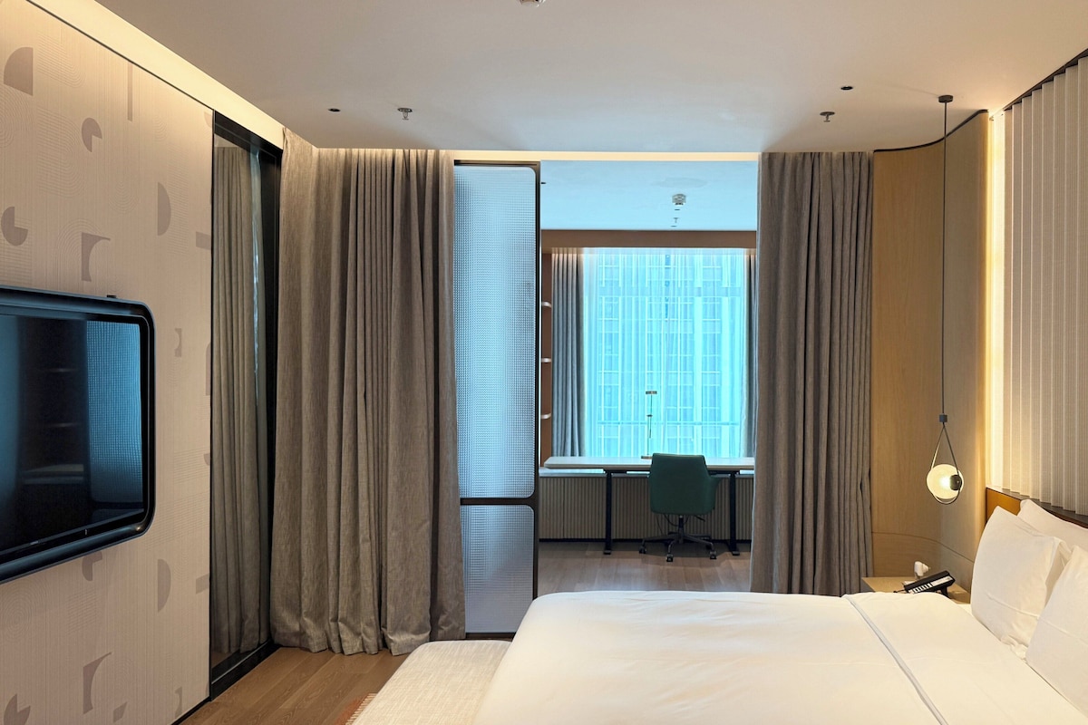 A hotel room with a large bed, a desk with a chair, a television, and a window with curtains.