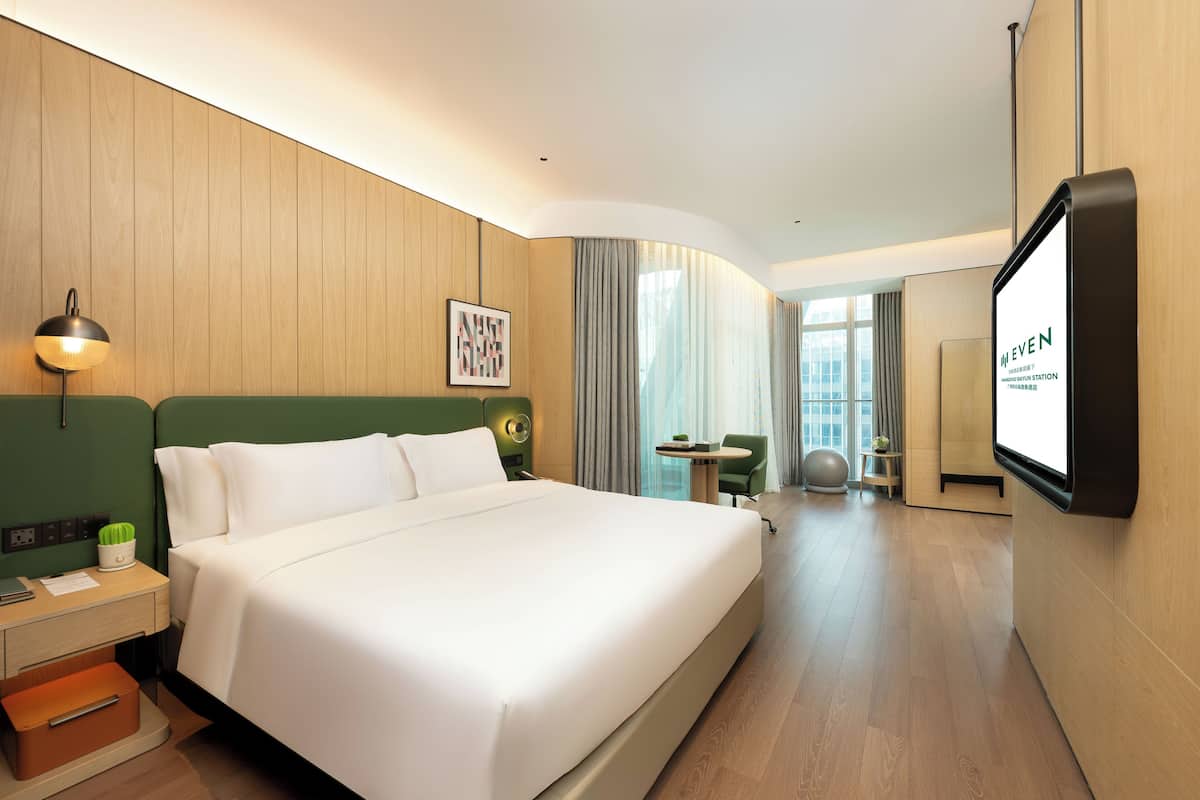 Habitación de hotel con una cama grande, un escritorio con lámpara, televisor y vistas a la ciudad a través de amplios ventanales.