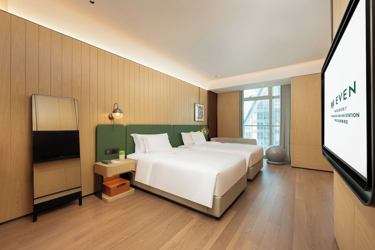 A hotel room with two beds, a flat-screen TV, a desk with a lamp, and a large window offering a view of the outside.