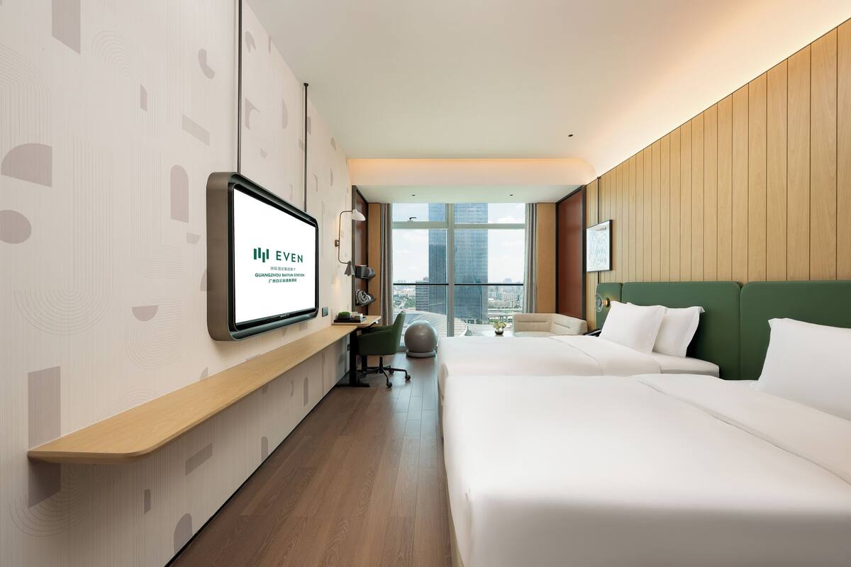 Habitación de hotel con una cama grande, un escritorio con computadora, una silla y vista a la ciudad a través de amplias ventanas.
